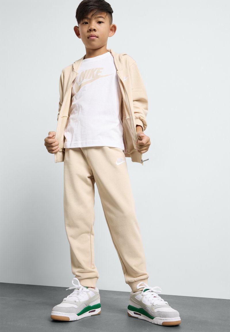 Bežinė hoodie ir treninginės kelnės su baltu Nike logotipu ant marškinėlių ir kelnių. Avalynė turi pilkus, baltus ir žalius akcentus.