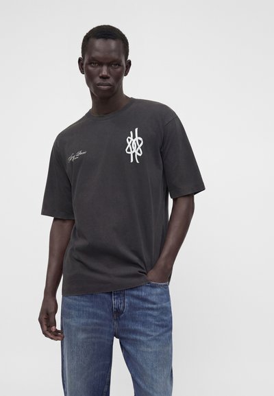 Άνδρας που φοράει ένα φαρδύ μαύρο T-shirt με λευκά γραφικά και κείμενο στο στήθος και μπλε τζιν, στέκεται με το ένα χέρι στην τσέπη.