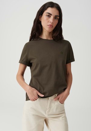 Vrouw met donker haar draagt een olijfgroen shirt met korte mouwen en lichtbeige broek, staand met handen in de zakken tegen een effen achtergrond.