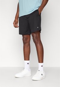 Μαύρο αθλητικό σορτς με ελαστική μέση, φορεμένο με λευκές κάλτσες και αθλητικά παπούτσια. Μπλε ανοιχτό πουκάμισο φορεμένο πάνω από το σορτς.