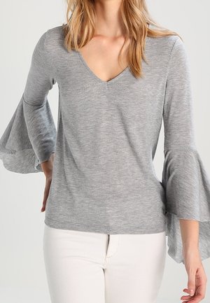 Lichtgrijs V-hals shirt met lange mouwen, voorzien van uitlopende doorschijnende klokmouwen en een gestructureerde stof, gecombineerd met een witte broek.