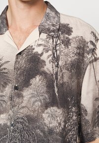 Camicia crema con stampa dettagliata di una foresta e alberi neri, maniche corte e bottoni neri nell'area superiore del petto e del collo di una persona.