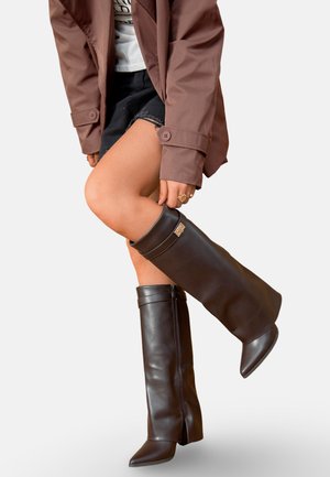 Personne portant des bottes en cuir brun foncé montant jusqu'aux genoux, un short noir, une chemise blanche et une veste marron, levant légèrement une jambe.
