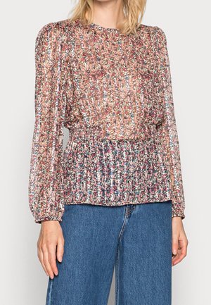 Femme portant un chemisier transparent à fleurs avec de longues manches bouffantes et une taille froncée, associé à un jean bleu taille haute.