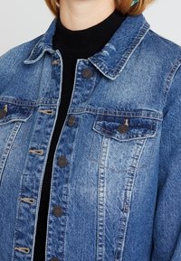 Džínová bunda s knoflíkovými náprsními kapsami, nošená přes černý žebrovaný rolák na osobě.