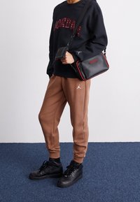 Felpa nera con scritta rossa "Jordan", pantaloni della tuta marroni con un logo bianco, sneakers nere e una piccola borsa nera con dettagli rossi.