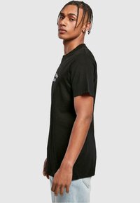 Black cotton t-shirt with short sleeves, crew neck, and printed text on the front; paired with light blue jeans, displayed in profile view.