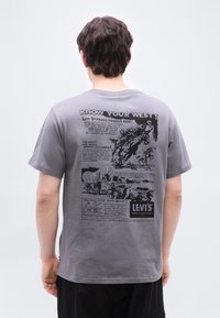 Άνδρας που φοράει γκρι μπλουζάκι με μαύρο τύπωμα θεματικό Δύσης της Levi's, που απεικονίζει καουμπόη, άλογα, καλυμμένα κάρα και διαφημιστικό κείμενο στην πλάτη.