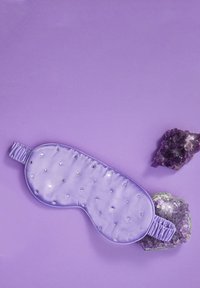 Mascherina per il sonno in satin lilla con piccoli borchiette argentate, forma sagomata e banda elastica arricciata, posizionata su uno sfondo viola con cristalli di ametista.