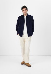Camisa de pana azul marino con botones y un único bolsillo en el pecho, combinada con pantalones blancos y zapatos marrones. Tela suave y texturada.