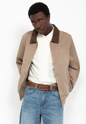 Mężczyzna w beżowej kurtce z ciemnym kołnierzem, białej koszuli na guziki, brązowym pasku i niebieskich dżinsach na białym tle.