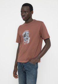 Καφέ βαμβακερό T-shirt με κοντά μανίκια, που διαθέτει ένα γραφικό σχέδιο τοπίου μέσα σε διακοσμητικό πλαίσιο στο μπροστινό μέρος.