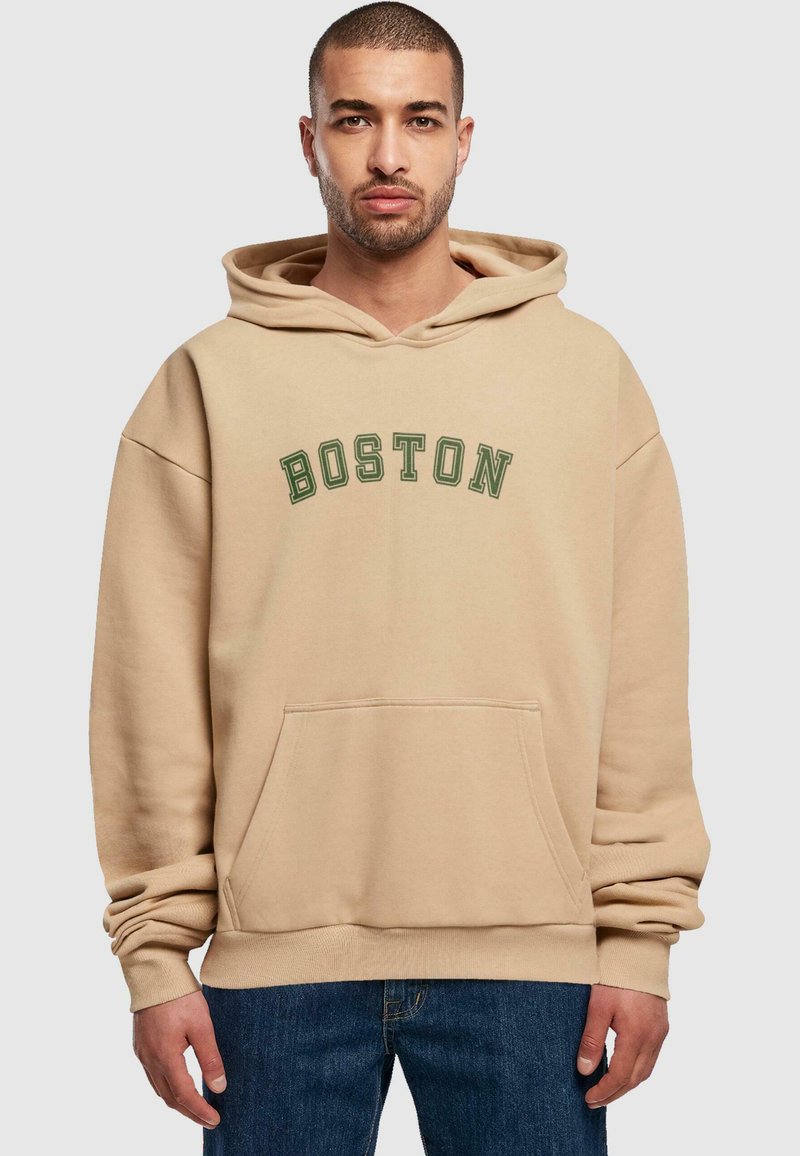 Beige kapucnis pulóver, zöld ívelt betűkkel "BOSTON" felirattal. Elöl zseb és bordázott mandzsetták találhatók rajta. Kék farmernadrággal viselve.
