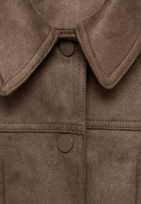 Braune Wildlederjacke mit strukturiertem Kragen, markanten Nähten und runden braunen Knöpfen. Texturierte Oberfläche mit subtiler Glanz.
