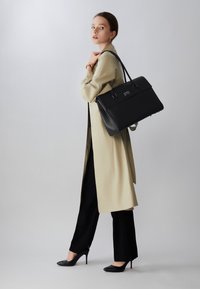Borsa nera strutturata con doppi dettagli a fibbia, superficie liscia e manici robusti, abbinata a un lungo trench coat beige e pantaloni neri.