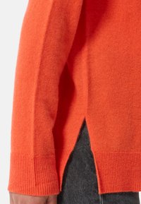 Maglione arancione in lana con polsini a costine e fessure laterali. Texture morbida e vestibilità rilassata, parzialmente mostrato con denim scuro sotto.