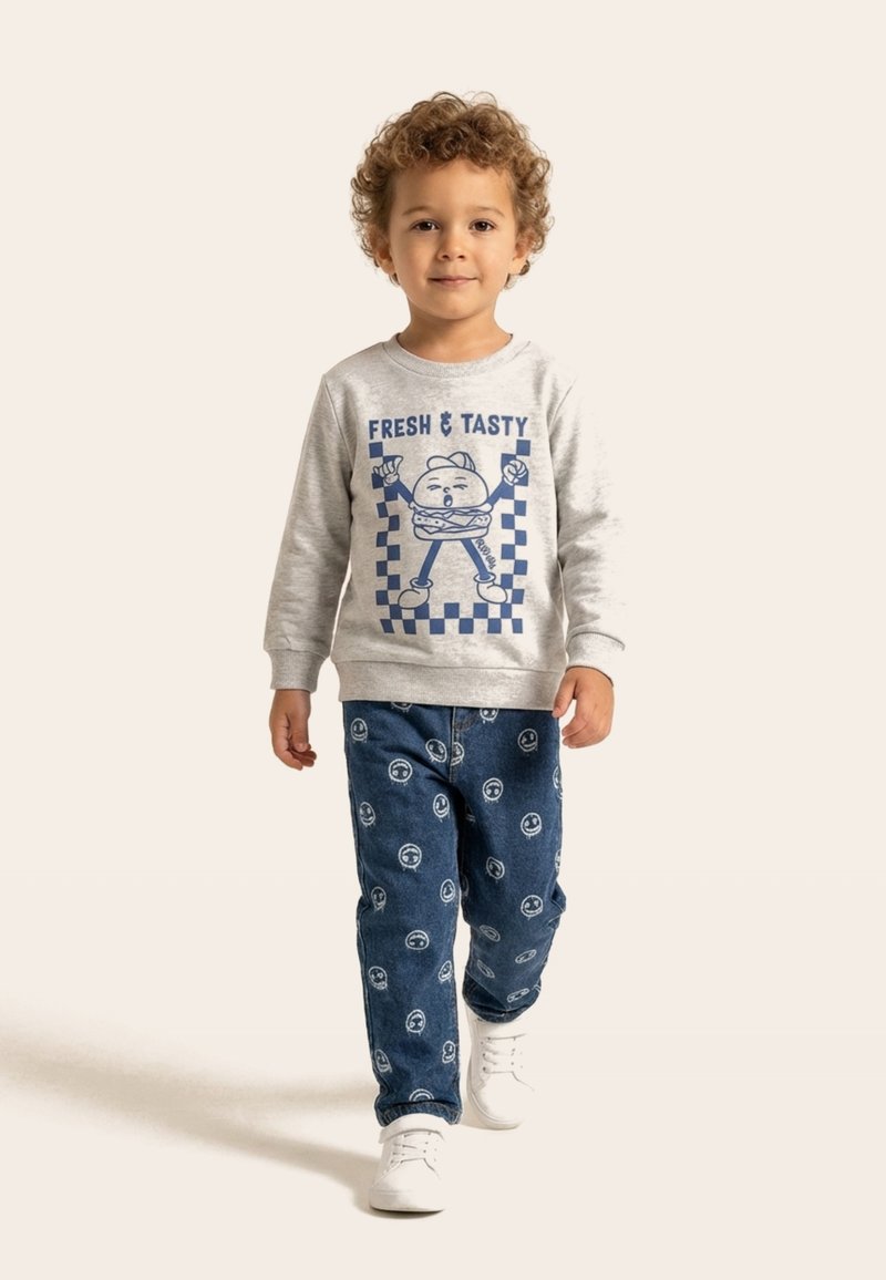 Copil mic cu păr creț purtând un hanorac gri cu grafică de burger și pantaloni albaștri cu fețe zâmbitoare albe, mergând înainte.