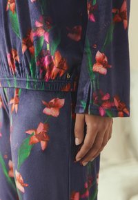 Lilleline koduriietuse komplekt tumesinisel taustal roosade ja roheliste orhideemustritega. Omadusteks on elastne vöökoht, pehme tekstuur ja vööl logoga aksent.