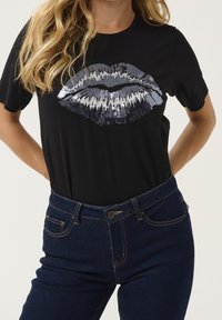 Černé tričko s velkým designem rtů z černých a stříbrných flitrů. Krátké rukávy a pohodlný volný střih, kombinované s modrými džínami.