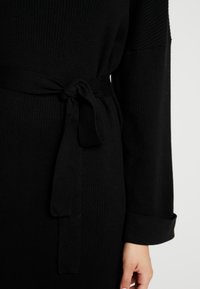 Zwarte ribgebreide jurk met een strikceintuur in de taille. Lange mouwen met opgerolde manchetten. Zachte textuur en aansluitend ontwerp.