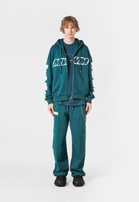 Sudadera color teal con gran texto gráfico blanco, combinada con pantalones de chándal a juego. Dispone de cordones, cremallera y una cadena decorativa. Tejido texturizado.