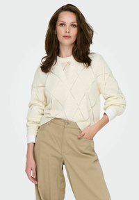 Femme aux cheveux bruns ondulés portant un pull en tricot crème avec des motifs en losanges et un pantalon beige ample, main dans la poche, sur fond clair.