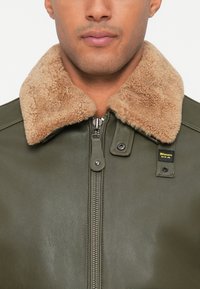 Uomo che indossa una giacca zip-up verde oliva con colletto in pile color beige e strap con bottone a pressione su uno sfondo bianco.