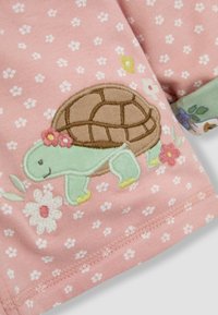 Tela rosa con bordado de una tortuga y acentos florales. La tortuga tiene un caparazón marrón y una expresión sonriente, sobre un fondo de puntos.