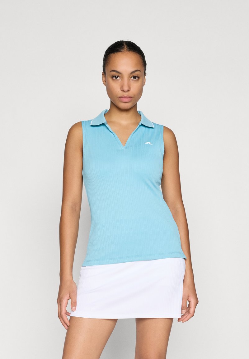 Camisola tipo polo sem mangas de cor azul clara com textura canelada e colarinho branco. Apresenta um pequeno logótipo do lado esquerdo. Usada com uma saia branca.