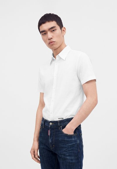 Jeune homme portant une chemise blanche à manches courtes boutonnée et un jean bleu foncé, une main dans la poche, sur un fond uni.