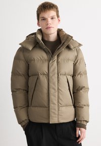 Khaki pufferjakke med tekstureret finish, isoleret design, stor hætte og forlommer. Kombineret med en ribbet brun sweater underneath.