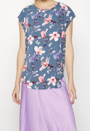 Femme portant un haut court bleu à motifs floraux avec des fleurs roses et violettes ainsi que des papillons, associé à une jupe violet clair.