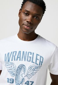 Camiseta de algodón blanca con el texto gráfico azul "WRANGLER" que incluye alas y un diseño de neumático, cuello redondo, mangas cortas y textura suave.
