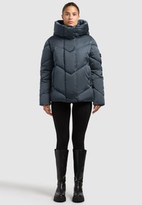 Tumesinist täidisega jope, millel on kõrge krae, struktureeritud ševroonmustritega ja elastsete mansettidega, kombineeritud mustade kitsaste retuuside ja põlveni ulatuvate saapadega.