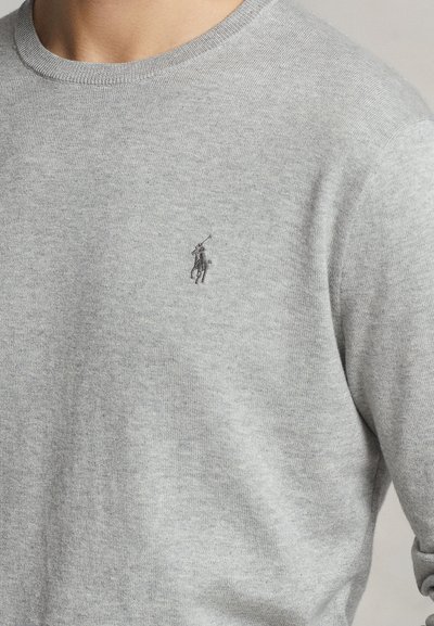 Primo piano di una persona che indossa un maglione grigio chiaro con un piccolo logo ricamato di un giocatore di polo sul petto sinistro.