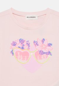 Ανοιχτό ροζ μπλουζάκι με σχέδιο γυαλιών ηλίου σε σχήμα καρδιάς με ροζ παγιέτες και ανθισμένες λεπτομέρειες. Η ετικέτα γράφει "BILLIEBLUSH".