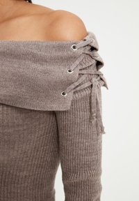 Pull à col bateau en maille côtelée marron clair avec détails à lacets sur les épaules, présentant des œillets métalliques et une texture douce.