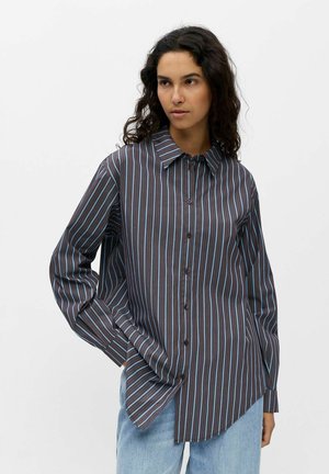 Femme aux cheveux bouclés foncés portant une chemise marron boutonnée avec des rayures verticales bleues et un jean bleu clair, debout devant un fond blanc.