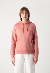 Felpa rosa realizzata in tessuto morbido, con tasca frontale, cappuccio con lacci regolabili e polsini a coste, abbinata a pantaloni bianchi.