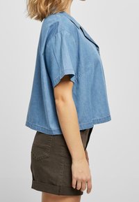 Woman wearing a loose, short-sleeve, light blue denim shirt and dark brown rolled-hem shorts, shown from the side against a plain background.