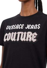 Must musta T-särk mustast puuvillast, millel on valge ja roosa rhinestone'e kaunistatud "COUTURE" logo ning sahistav taust allpool "VERSACE JEANS".