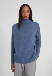 Donna con capelli ricci che indossa un maglione a collo alto blu e pantaloni grigio chiaro, in piedi davanti a uno sfondo semplice e chiaro.