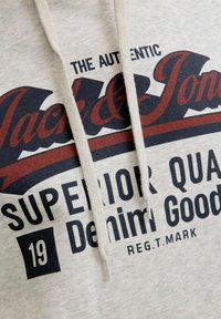 Světle šedá mikina s kapucí s velkým, výrazným potiskem v námořnické modři a burgundské barvě, s tkaničkami, na kterém je nápis "Jack & Jones" a "Superior Quality Denim Goods."