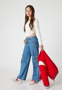Chaqueta de varsity roja con acentos en blanco y azul marino, top corto de manga larga blanco, pantalones cargo holgados azules y zapatillas grises con detalles rojos.