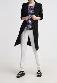 Blazer lungo nero sopra una blusa floreale blu navy, pantaloni bianchi slim-fit e sandali neri con cinturini. Tessuto liscio, forma sartoriale, design moderno.