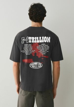 Fekete pólót viselő személy, amelyen nagy fehér felirat olvasható: "F-1 Trillion", piros grafika, dalcímek listája és a "Posty Co." logó a hátoldalon.