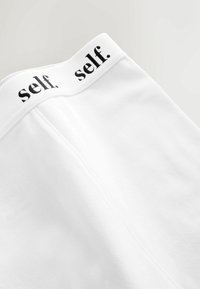 Valkoiset kangashousut, joissa on pehmeä pinta, varustettu mustalla elastisella vyötärönauhalla, jossa on toistuva teksti "self.".