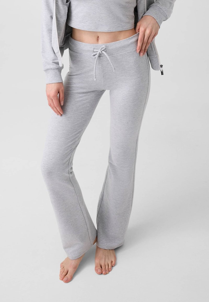 Femme portant un pantalon de survêtement gris clair avec cordon de serrage et un top court ainsi qu'une veste assortis, debout pieds nus sur un fond clair.