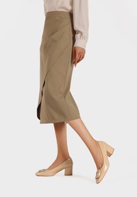 Gonna midi khaki con dettaglio drappeggiato diagonale, abbinata a scarpe con tacco grosso in pelle verniciata beige con una fibbia decorativa.