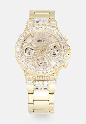 Montre-bracelet Guess dorée avec accents en cristal, chiffres romains, trois sous-cadrans et un bracelet métallique à maillons.